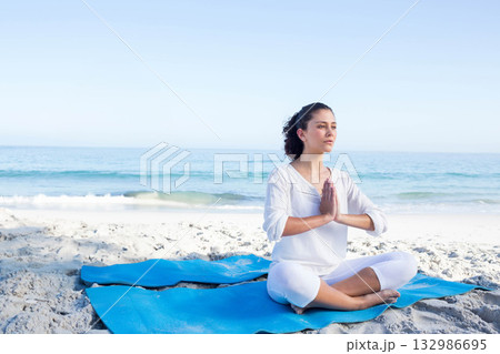 Woman meditating on blue yoga mat on beach wearing white activewear holding palms, copy space 132986695