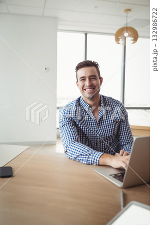 Smiling man wearing checkered shirt working at wooden desk at office using laptop and smartphone 132986722