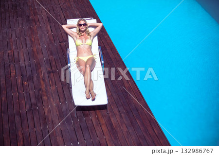 Woman reclining wearing yellow bikini and sunglasses on white lounge chair on deck beside blue pool 132986767