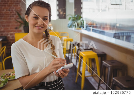 Female server holding order pad and pen inside café beside table with salad wearing striped apron Female server holding order pad and pen inside café beside table with salad wearing striped apron 132986776