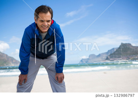 Senior man catching breath with hands on knees on beach wearing blue windbreaker and grey pants 132986797