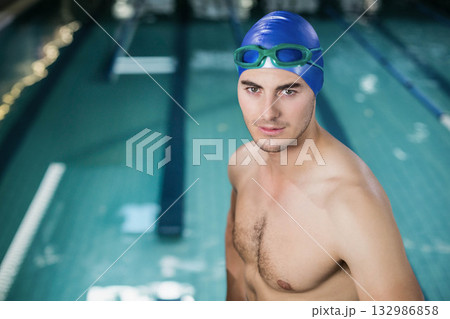 Male swimmer standing at pool edge wearing blue cap, green goggles by lane ropes, copy space 132986858