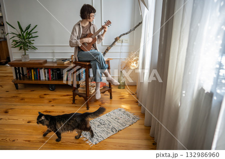 Focused young woman rehearsing ukulele by window, immersed in chord repetition with cat walk around Focused young woman rehearsing ukulele by window, immersed in chord repetition with cat walk around 132986960