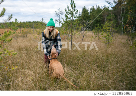 Happy woman bend to stroke dog neck in tall grassfield, canine look upward playing with owner Happy woman bend to stroke dog neck in tall grassfield, canine look upward playing with owner 132986965