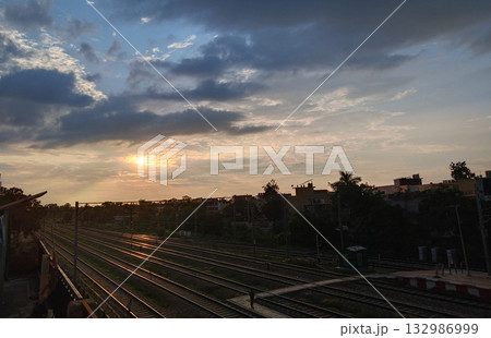 Cloudy evening sky with sun setting over railway lines and silhouetted buildings in the distance 132986999