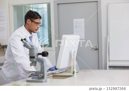 Male researcher wearing lab coat, gloves typing on PC at lab bench with microscope, pipette stand 132987268