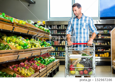 Mid adult man pushing shopping cart of produce by wooden bins in supermarket aisle, copy space 132987286
