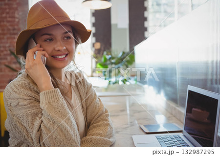 Woman wearing fedora calling on smartphone near laptop and tablet at cafe counter, copy space 132987295