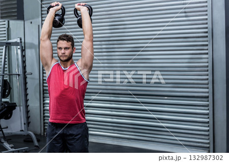 Male athlete in red shirt lifting kettlebells at gym with weight rack and metal shutter door 132987302