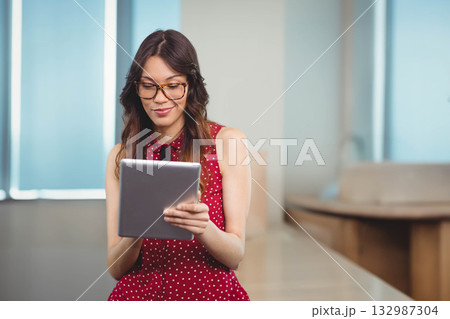 Woman wearing eyeglasses holding tablet and reading screen in office conference area with blinds 132987304