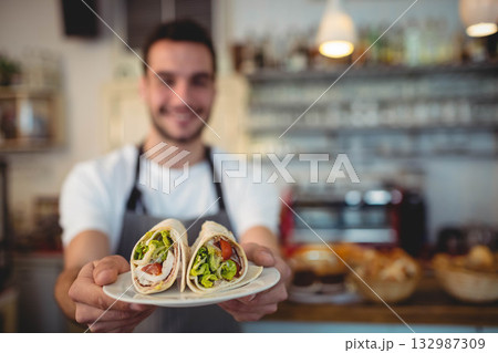 Barista wearing apron holding two tortilla wraps with lettuce and tomato on plate at café counter 132987309