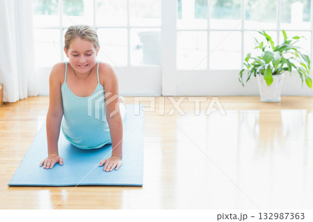 Female child practicing yoga backbend on blue mat on wood floor beside glass doors, copy space 132987363