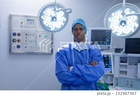 Mid adult male surgeon wearing blue surgical gown in operating room under lights, watching monitor Mid adult male surgeon wearing blue surgical gown in operating room under lights, watching monitor 132987367