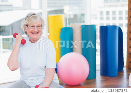 Senior female smiling and lifting red dumbbell by shoulder on bench in fitness studio, copy space Senior female smiling and lifting red dumbbell by shoulder on bench in fitness studio, copy space 132987371