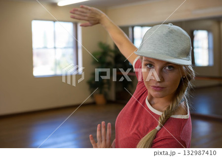 Female dancer wearing gray cap practicing pose by ballet barre before mirrored wall at dance studio 132987410