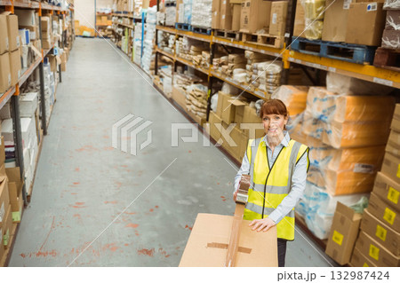 Female warehouse worker applying packing tape onto box in warehouse aisle with shelving, copy space 132987424