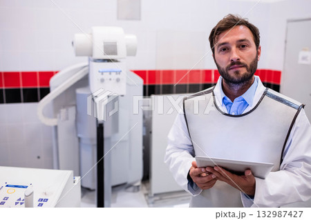 Male radiology technician wearing lead apron at x-ray machine holding tablet in clinic, copy space Male radiology technician wearing lead apron at x-ray machine holding tablet in clinic, copy space 132987427