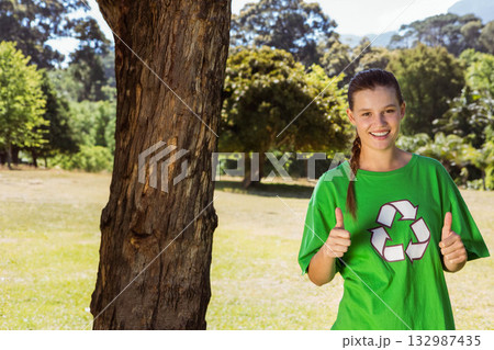 Woman standing next to tree trunk in sunny meadow wearing green recycling shirt, copy space Woman standing next to tree trunk in sunny meadow wearing green recycling shirt, copy space 132987435