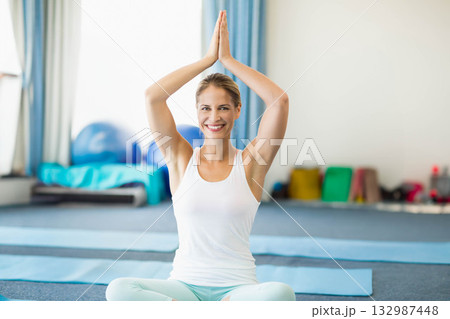Adult woman smiling, performing seated yoga pose on yoga mat in yoga studio near exercise balls Adult woman smiling, performing seated yoga pose on yoga mat in yoga studio near exercise balls 132987448
