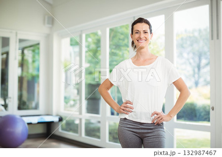 Woman standing beside purple stability ball and bench in fitness room by large windows, copy space Woman standing beside purple stability ball and bench in fitness room by large windows, copy space 132987467