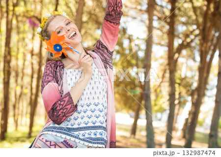 Woman posing in forest park wearing floral headband holding orange pinwheel raising arm 132987474