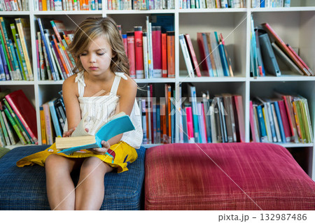 School-age girl sitting on dark blue and red ottomans, turning page of turquoise book in library 132987486