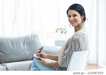 Woman writing notes on clipboard with pen sitting sideways on white chair in bright home office 132987505