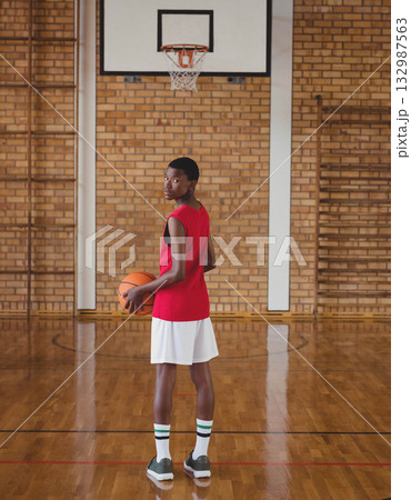 Chinese teenage boy wearing red jersey holding basketball under school gym hoop and looking back 132987563
