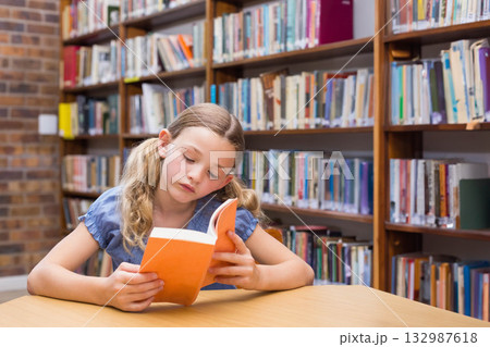 Female child reading orange-covered paperback at library table near brick wall, bookshelves 132987618