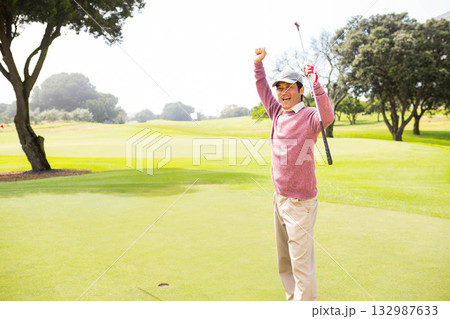 Asian mid adult man celebrating putt on golf green holding putter and flagstick, copy space 132987633