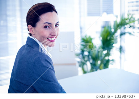 Businesswoman in suit smiling at camera, turning at office desk with horizontal blinds and plant 132987637