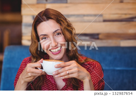 Middle-aged woman in red blouse smiling and holding latte on blue bench in coffee shop Middle-aged woman in red blouse smiling and holding latte on blue bench in coffee shop 132987654
