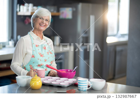 Senior woman pouring cupcake batter into muffin tin on countertop wearing floral apron, copy space 132987667