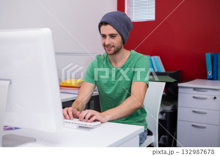Man wearing gray beanie and green shirt typing on compact keyboard at white desk in office Man wearing gray beanie and green shirt typing on compact keyboard at white desk in office 132987678