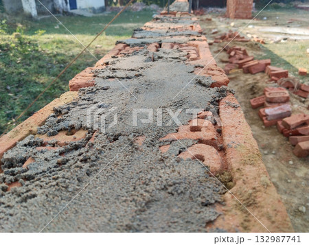 Closeup view of bricklaying process with cement mortar applied on red bricks for boundary wall construction in rural area, traditional masonry technique in Indian village construction work 132987741