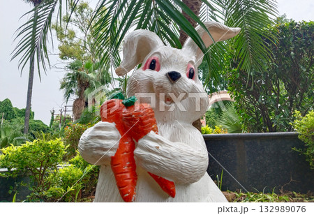 Cute white rabbit statue holding carrots in park garden, decorative animal figure among greenery, playful attraction for children Cute white rabbit statue holding carrots in park garden, decorative animal figure among greenery, playful attraction for children 132989076