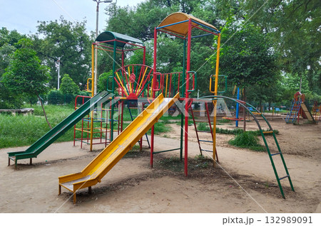 Colorful metal playground structure with ladders, yellow and green slides, climbing arches, and shaded platforms in outdoor park setting for children 132989091