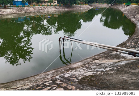 Water canal pond with stone embankment and metal pipes, outdoor artificial reservoir in public park used for irrigation and water storage surrounded by greenery and reflecting trees in calm surface Water canal pond with stone embankment and metal pipes, outdoor artificial reservoir in public park used for irrigation and water storage surrounded by greenery and reflecting trees in calm surface 132989093