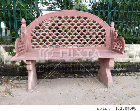 Ornate pink stone bench with traditional lattice-pattern backrest and decorative armrests, installed outdoors in park setting, blending heritage architecture with public seating for relaxation Ornate pink stone bench with traditional lattice-pattern backrest and decorative armrests, installed outdoors in park setting, blending heritage architecture with public seating for relaxation 132989099