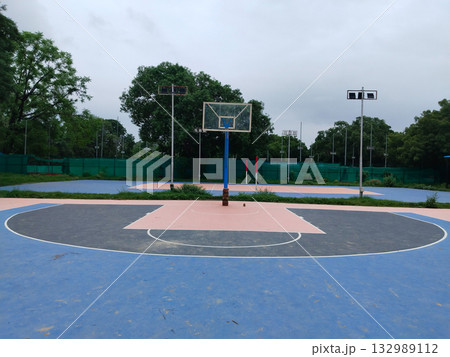 Empty outdoor basketball court with pink and blue painted surface, lone hoop post in quiet sports complex surrounded by greenery and tall light poles, peaceful athletic ground for games and recreation 132989112