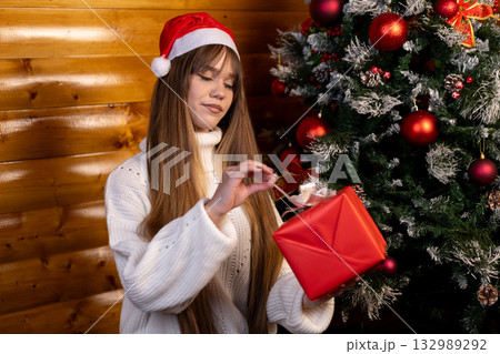 Young girl in a Santa hat opens a present box by a Christmas tree 132989292