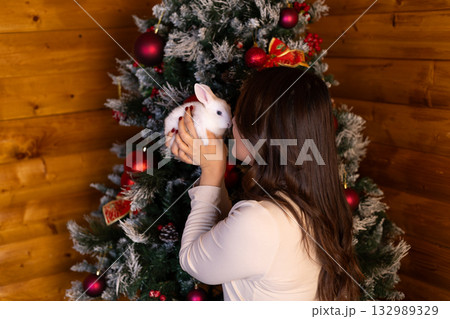 A woman is holding a rabbit and nuzzling it next to a decorated Christmas tree. 132989329