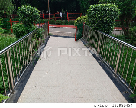Tiled pedestrian ramp with stainless steel railings and green plants, leading to gate and road in public park or garden setting Tiled pedestrian ramp with stainless steel railings and green plants, leading to gate and road in public park or garden setting 132989454
