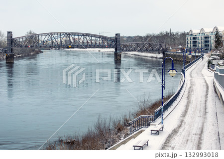 Snowy morning at Magdeburg Schleinufer shows calm river Elbe reflecting steel Hubbrucke bridge. Quiet winter path lined with blue lamps and benches creates serene urban landscape 132993018