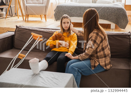 Girl with broken leg in cast sits with mother on couch at home. Sad little kid hugging teddy bear recovering from broken leg. Girl using cast crutches for quick rehabilitation. Mom supporting daughter 132994445