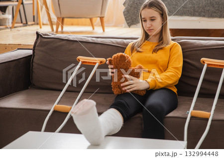 Young girl with broken leg cast sitting on sofa. Sad little kid hugging her teddy bear recovering from broken leg at home. Girl using cast and crutches. Accident injury fracture treatment foot bandage 132994448