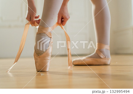 Ballerina puts on pointe shoes on leg in dance class. Classical ballet dancer woman in dance studio. Ballerina putting on tying classical pointe shoes for dance training. Ballet school concept 132995494