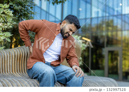 A man seated outdoors on a wooden bench, displaying discomfort due to back pain. The setting includes a modern building and greenery, conveying a combination of pain and concern A man seated outdoors on a wooden bench, displaying discomfort due to back pain. The setting includes a modern building and greenery, conveying a combination of pain and concern 132997528
