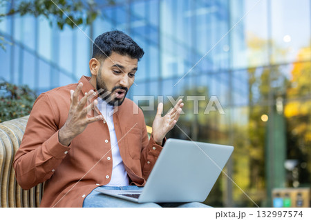 Shocked businessman is reacting to bad news on his laptop while sitting on a bench outdoors, expressing disbelief and frustration with raised hands 132997574