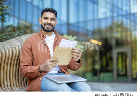 Happy young man sitting outdoors on a modern bench, smiling and holding an open envelope with an acceptance letter, possibly receiving good news about his university admission 132997613